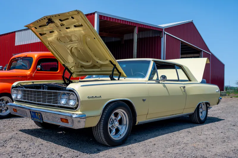 Cream two-door muscle car with its hood raised, parked three-quarter front on gravel beside a red barn