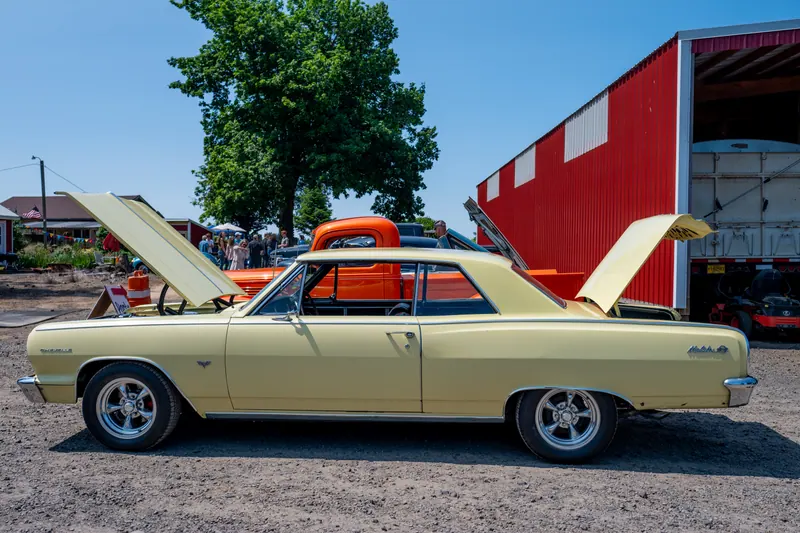 The same cream muscle car in side profile with hood and trunk open, show-ready on gravel