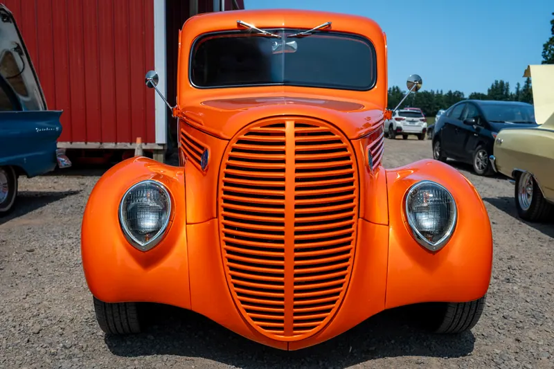 Front end of a bright orange hot rod pickup, chrome grille and headlights filling the frame against red barn siding