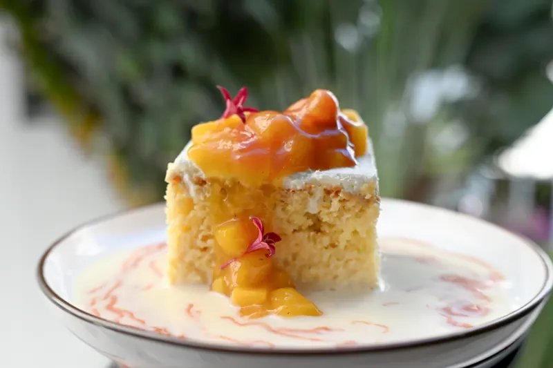 Slice of tres leches cake topped with caramel and diced mango, plated with a red sauce swirl