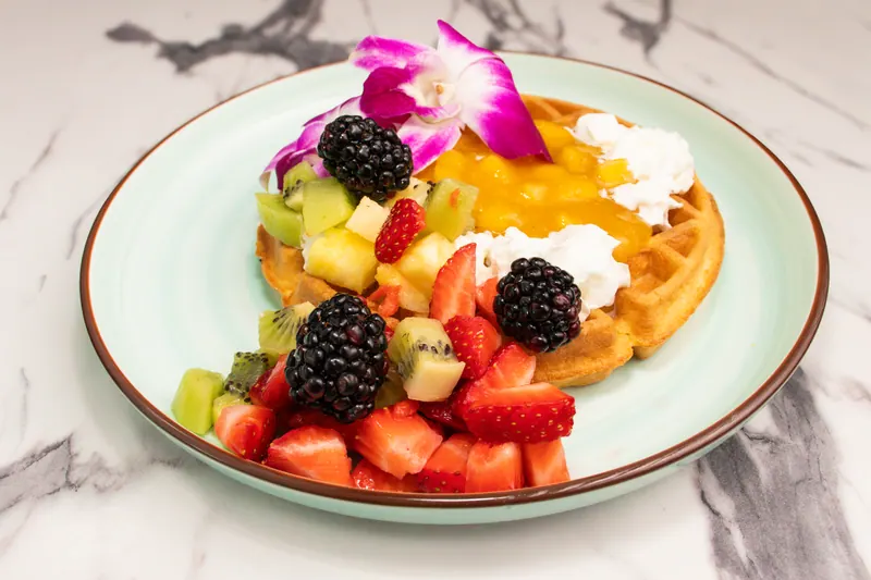 Belgian waffle topped with strawberries, blackberries, kiwi and mango, garnished with an orchid, on a pale plate