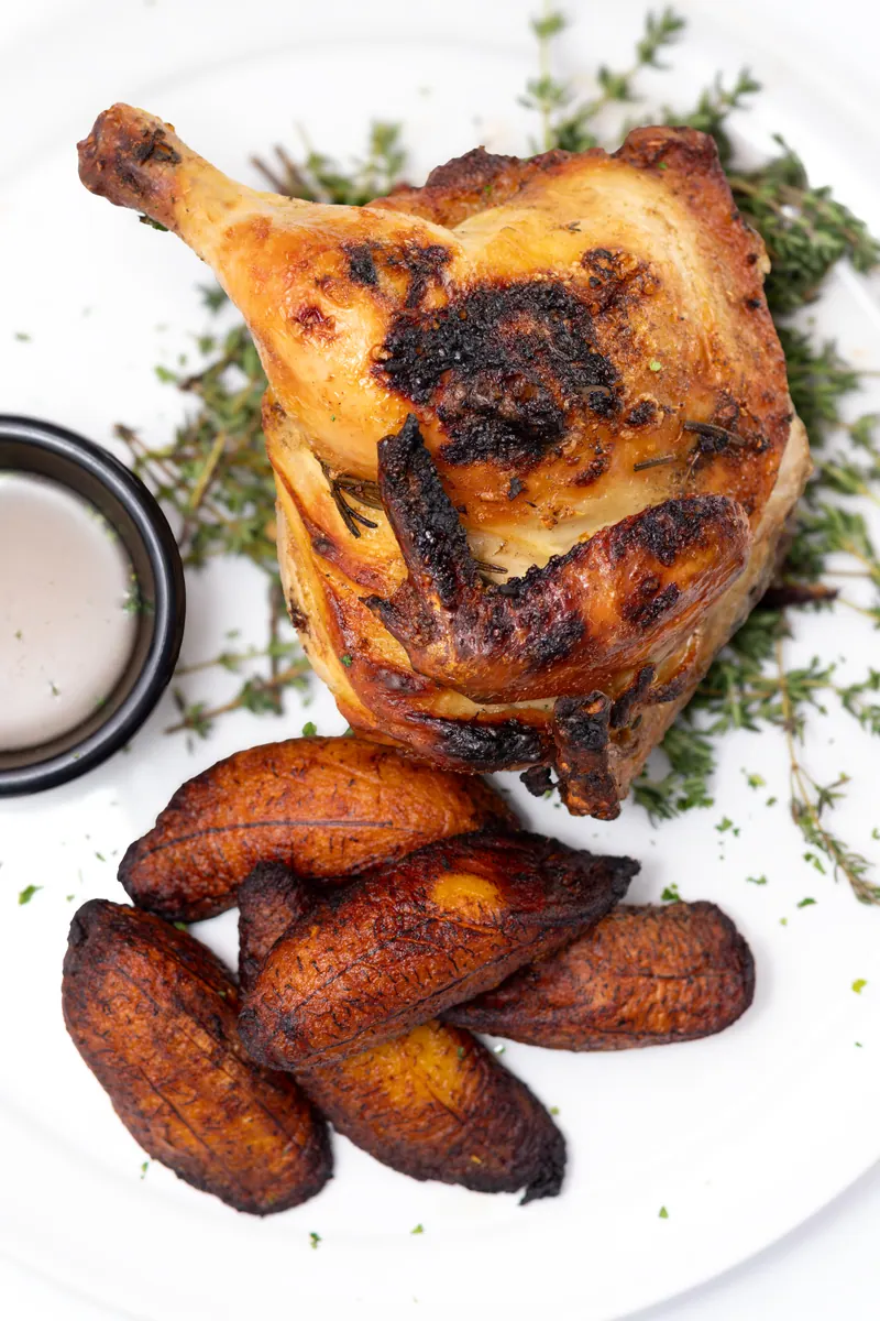 Roast half chicken with charred skin alongside caramelised sweet plantains, shot against a bright outdoor background